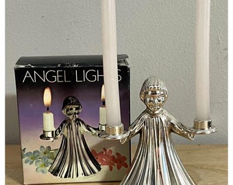 Puede incluir: Un candelabro de metal plateado con forma de ángel con los brazos extendidos que sostiene dos velas cónicas blancas delgadas. El producto se muestra junto a su caja original negra y morada con la etiqueta ANGEL LIGHTS.