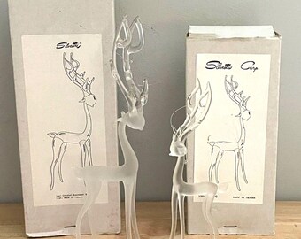 Puede incluir: Dos figuras de reno de cristal esmerilado transparente con astas altas y ramificadas. Cada una se sitúa frente a su caja de cartón rectangular original, que presenta un dibujo lineal negro del reno y el nombre de marca Silvestri.