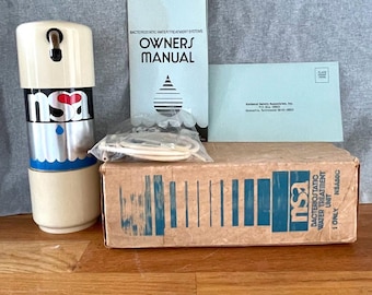 Puede incluir: Una unidad de tratamiento de agua NSA cilíndrica de color blanco roto y plateado con base azul, acompañada de un manual de usuario, una manguera blanca envuelta en plástico y un sobre de National Safety Associates junto a una caja de cartón.