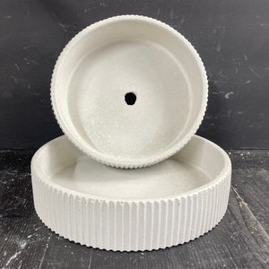 May include: Two white concrete planters with a ribbed design. The top planter has a drainage hole in the center.