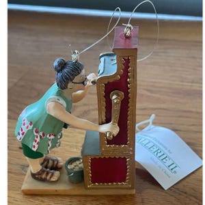 May include: A decorative ornament featuring a figure with gray hair in a bun, wearing a green dress with red and white patterns, standing at a red and gold slot machine. The figure holds the machine's handle. A small bucket sits at the base.