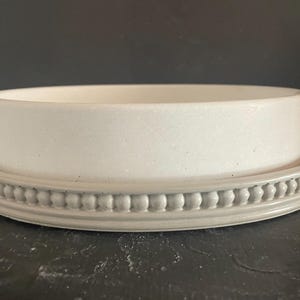 May include: A white ceramic planter with a matching saucer. The planter is cylindrical and sits inside the saucer. The saucer has a decorative beaded edge. The items are a light gray color.
