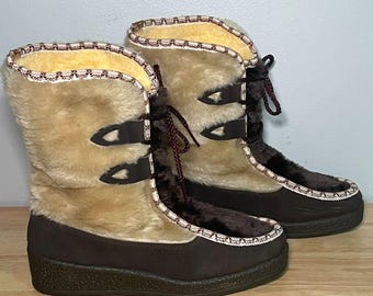 Puede incluir: Un par de botas de invierno de media caña con secciones inferiores en marrón oscuro y partes superiores de piel sintética beige. Las botas incluyen cordones marrón oscuro con pestañas decorativas, suela de goma negra texturizada y ribete.
