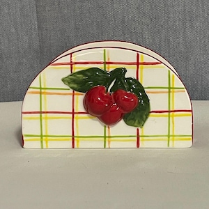 May include: Ceramic napkin holder with a white background and a colorful plaid pattern of red, yellow, and green lines. The holder features a 3D cherry design with two red cherries and green leaves.