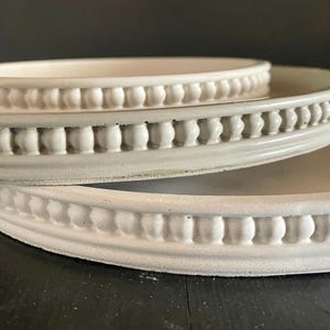 May include: Three off-white ceramic trays of varying sizes are stacked. The trays have a raised, beaded edge design. The largest tray is at the bottom, with a medium-sized tray in the middle and a small tray on top. The trays are on a dark surface.