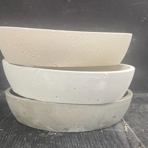 May include: Three concrete planters in different sizes, stacked on top of each other. The top planter is a light beige color, the middle planter is white, and the bottom planter is a light gray color.