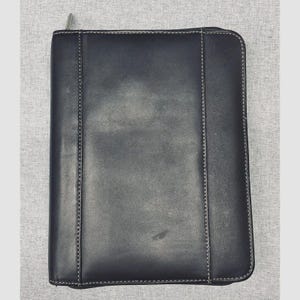 May include: A rectangular black leather portfolio case featuring a smooth surface, rounded corners, and visible white stitching along the vertical panels. The case includes a silver-toned metal zipper pull located at the top left corner.