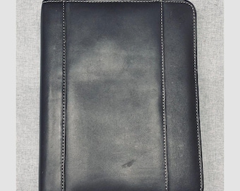 Puede incluir: Una carpeta rectangular de cuero negro con superficie lisa, esquinas redondeadas y costuras blancas visibles a lo largo de los paneles verticales. El estuche incluye un tirador de cremallera de metal plateado en la esquina superior izquierda.
