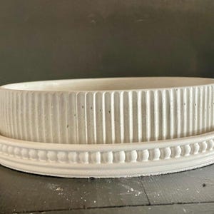 May include: A white, textured, concrete planter with a matching saucer. The planter has vertical ribbed detailing on the exterior, while the saucer features a decorative beaded edge. This item is suitable for indoor or outdoor use.