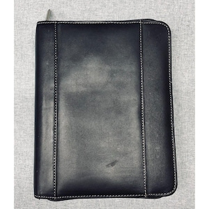 May include: A rectangular black leather portfolio case featuring a smooth finish and white contrast stitching along the vertical panels. The case includes a silver metal zipper pull located at the top left corner, providing a secure closure.