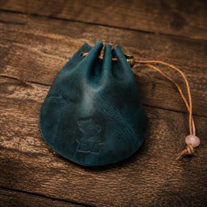 May include: A teal leather drawstring pouch featuring a debossed anvil logo on the front. The pouch is cinched with a thin tan leather cord that includes a small metallic bead and a light pink stone bead at the end of the cord strings.
