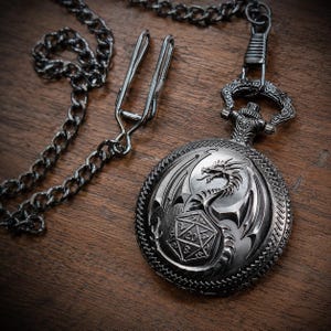 May include: A silver-coloured pocket watch featuring an embossed dragon design on the front cover, which also displays a twenty-sided die with the number 20 visible. The watch is attached to a matching silver-coloured metal chain with a belt clip.