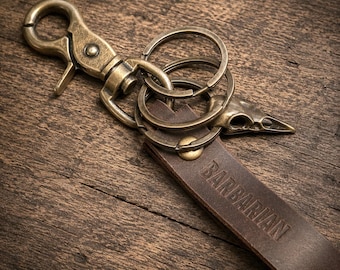 Könnte beinhalten: Ein dunkelbrauner Schlüsselanhänger aus Leder mit dem eingeprägten Wort BARBARIAN auf der Oberfläche. Das Band ist an einem messingfarbenen Karabinerhaken, zwei runden Spaltringen und einem kleinen Metallanhänger in Form eines Vogelschädels befestigt.