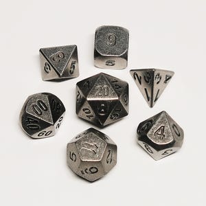 Puede incluir: Un conjunto de siete dados metálicos en varias formas geométricas, incluyendo un d20, d12, d10, d8, d6 y d4. Los dados son de color gris plateado con números grabados, para juegos de rol.