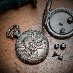 Puede incluir: Una caja de reloj de bolsillo de metal plateado con un diseño en relieve de un pulpo y un ojo en la tapa. La caja está abierta, revelando un interior liso, y está acompañada por cuatro dados de juego oscuros de varias caras cercanos.