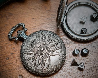 May include: A silver-toned metal pocket watch case featuring an embossed octopus and eye design on the cover. The case is open, revealing a smooth interior, and is accompanied by four small, dark, multi-sided gaming dice scattered nearby.