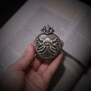 K&ouml;nnte beinhalten: Eine runde, bronzefarbene Taschenuhr mit einem komplizierten Pr&auml;gemuster, das ein zentrales Auge umgeben von mehreren Tentakeln zeigt. Die Uhr hat eine dekorative Krone an der Oberseite und ist an einer passenden Metallkette befestigt.