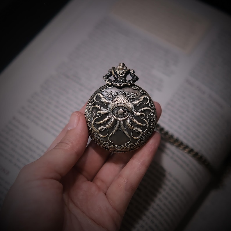 K&ouml;nnte beinhalten: Eine runde, bronzefarbene Taschenuhr mit einem komplizierten Pr&auml;gemuster, das ein zentrales Auge umgeben von mehreren Tentakeln zeigt. Die Uhr hat eine dekorative Krone an der Oberseite und ist an einer passenden Metallkette befestigt.