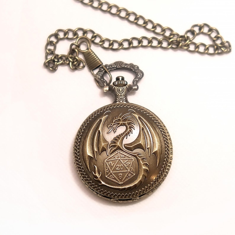 K&ouml;nnte beinhalten: Eine runde Taschenuhr in antikem Bronzeton mit einem gepr&auml;gten Drachenmotiv, das sich um einen zwanzigseitigen W&uuml;rfel windet. Die Uhr ist an einer passenden bronzefarbenen Metallkette befestigt, wobei der W&uuml;rfel deutlich die Zahl 20 oben zeigt.