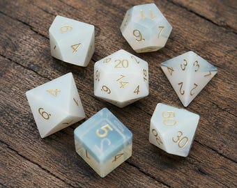 May include: A set of seven polyhedral dice made of a semi-translucent, milky white material with a subtle blue tint. Each die features engraved numbers filled with gold-colored ink, including a twenty-sided die, a twelve-sided die, and others.