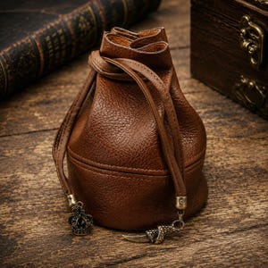 Pode incluir: Uma bolsa de couro castanho texturizado com fecho de cordão e base arredondada. O fecho possui dois cordões de couro, cada um terminado com um pendente metálico: um em forma de pequena coroa com pedra escura e outro em forma de garra curva.