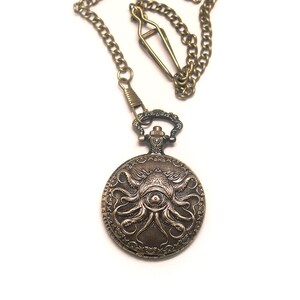 K&ouml;nnte beinhalten: Eine bronzefarbene Taschenuhr mit einem gepr&auml;gten Design eines zentralen Auges, das von Tintenfisch-Tentakeln auf dem Deckel umgeben ist. Die Uhr ist an einer passenden bronzefarbenen Metallkette mit einem Verschluss am Ende befestigt.
