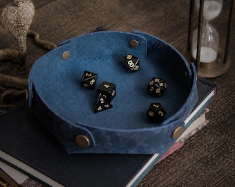 Op de afbeelding: Een blauwe lederen dobbelsteenbak met een zeshoekige vorm, voorzien van messingkleurige metalen drukknopen op elke hoek. In de bak liggen vijf zwarte veelzijdige dobbelstenen met goudkleurige cijfers, waaronder een d20, d12, d10, d8 en d4.
