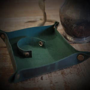 Pode incluir: Uma bandeja quadrada de couro verde-azulado com uma alça curva e fecho de pressão. A bandeja tem cantos arredondados e fechos de pressão cor de latão. O fundo inclui um vaso desfocado e uma superfície de madeira.
