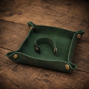 May include: A square dark green leather valet tray featuring four brass-coloured metal snaps at the corners to create raised edges. A small, arched leather loop is secured to the centre of the tray base with two matching brass-coloured metal snaps.