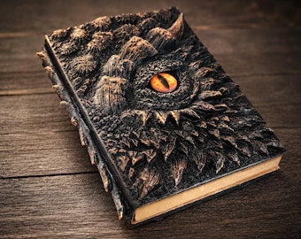 May include: A rectangular journal with a dark, textured cover featuring a sculpted dragon eye with a bright orange iris and black pupil. The cover has raised, scale-like details in black and bronze tones, and the pages have a light tan edge.