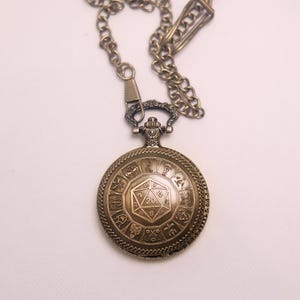 K&ouml;nnte beinhalten: Eine bronzefarbene Taschenuhr mit einem zwanzigseitigen W&uuml;rfeldesign auf dem Deckel, umgeben von zw&ouml;lf kreisf&ouml;rmigen Symbolen im Tierkreiszeichen-Stil. Die Uhr ist an einer passenden bronzefarbenen Metallkette mit einem sicheren Verschluss befestigt.