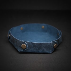 Puede incluir: Una bandeja de cuero azul con forma hexagonal. La bandeja tiene un borde elevado y está asegurada con remaches de color latón. El cuero tiene un aspecto envejecido. Ideal para organizar objetos pequeños, como llaves o joyas.