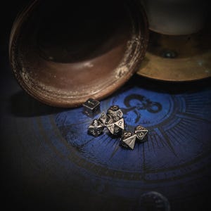 May include: A set of seven metallic, gunmetal-colored polyhedral dice featuring engraved black numbers. The dice set includes a d20, d12, d10, d8, d6, d4, and a percentile die, all arranged in a cluster on a dark blue surface with circular patterns.