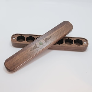 May include: Two wooden dice boxes with a dark brown finish. Each box has a series of hexagonal cutouts and a Celtic knot design. One box has a small, round, green stone. The boxes are designed to hold dice.