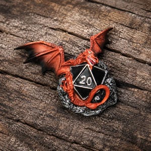 May include: A three-dimensional pin featuring a red dragon with black-tipped wings perched atop a black twenty-sided die. The die displays the number 20 in white. The dragon rests on a textured, silver-colored rocky base with intricate details.