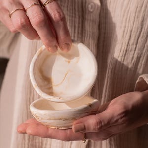 Op de afbeelding: Twee kleine, ronde, gebroken witte kommen met een gemarmerd, goudbruin patroon. De kommen worden in de handen van een persoon gehouden, met verschillende gouden ringen aan de vingers. De kommen zijn op elkaar gestapeld.