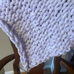 May include: A soft, fluffy, lavender-colored knitted blanket with a white yarn woven throughout. The blanket is draped over a wooden chair.
