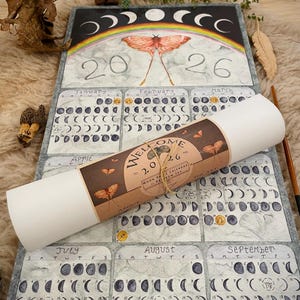 Puede incluir: Un calendario de fases lunares de 2026 con un dise&ntilde;o de acuarela. El calendario presenta un arco&iacute;ris, una polilla y las fases de la luna. Un rollo de papel con texto est&aacute; encima del calendario.