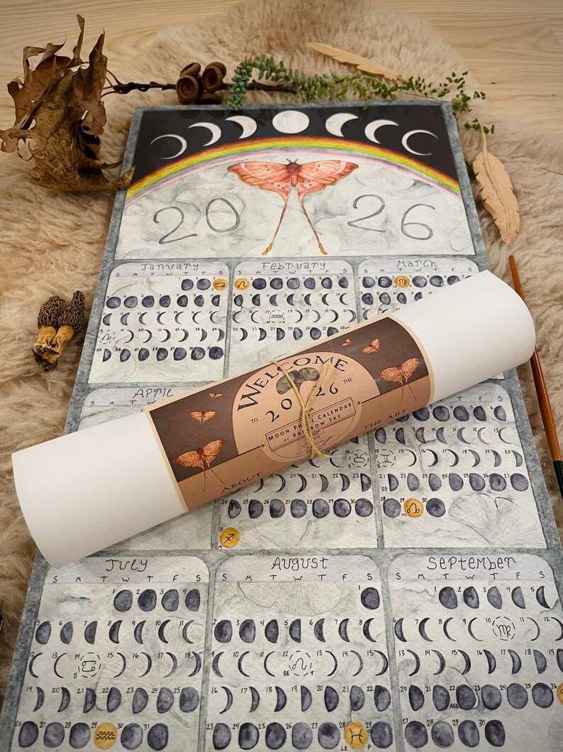 Puede incluir: Un calendario de fases lunares de 2026 con un dise&ntilde;o de acuarela. El calendario presenta un arco&iacute;ris, una polilla y las fases de la luna. Un rollo de papel con texto est&aacute; encima del calendario.