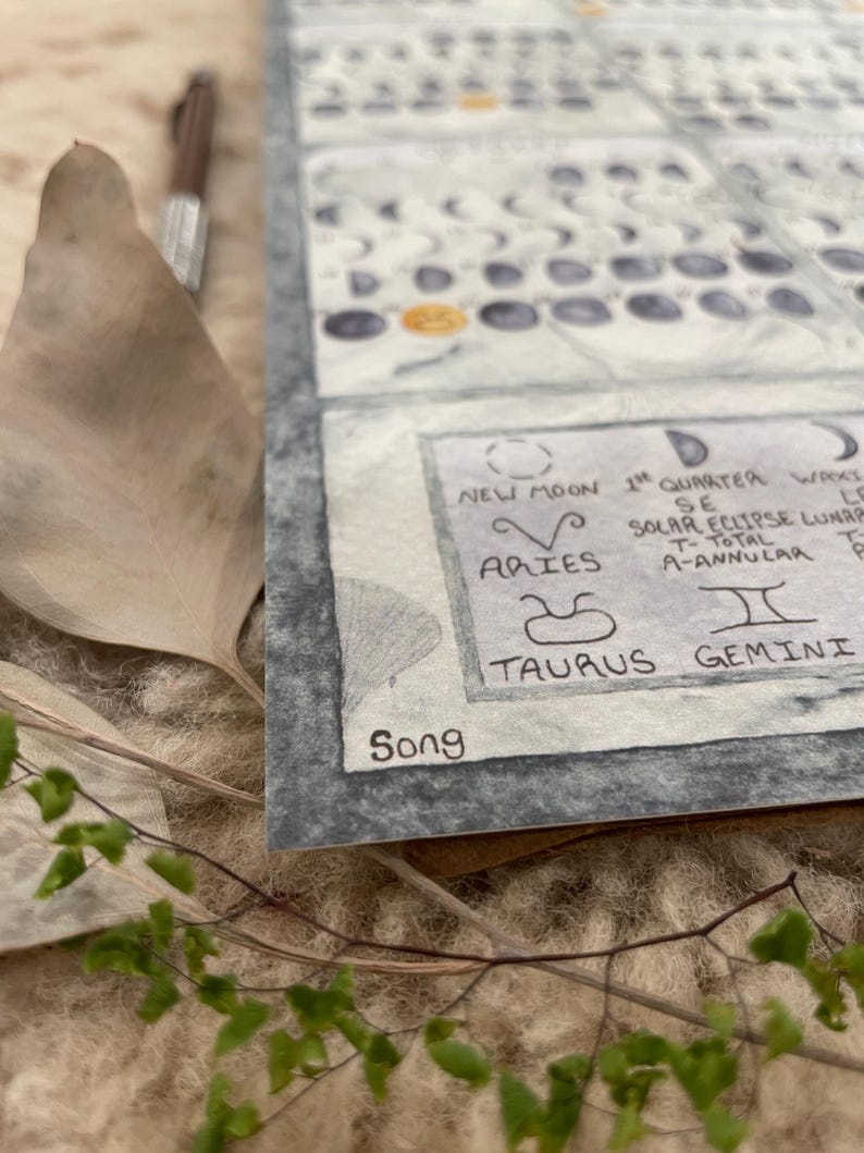 Puede incluir: Un calendario detallado en acuarela con s&iacute;mbolos astrol&oacute;gicos y fases lunares. El calendario incluye el texto "Luna Nueva", "1er Cuarto", "Aries", "Tauro" y "G&eacute;minis". La firma del artista "Song" es visible. Un bol&iacute;grafo y hojas secas est&aacute;n en el fondo.