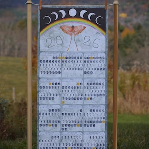 Puede incluir: Calendario de 2026 con una ilustraci&oacute;n de polilla, arco&iacute;ris y fases lunares. El calendario se muestra en un soporte de madera al aire libre, con meses y fechas dispuestas en una cuadr&iacute;cula.