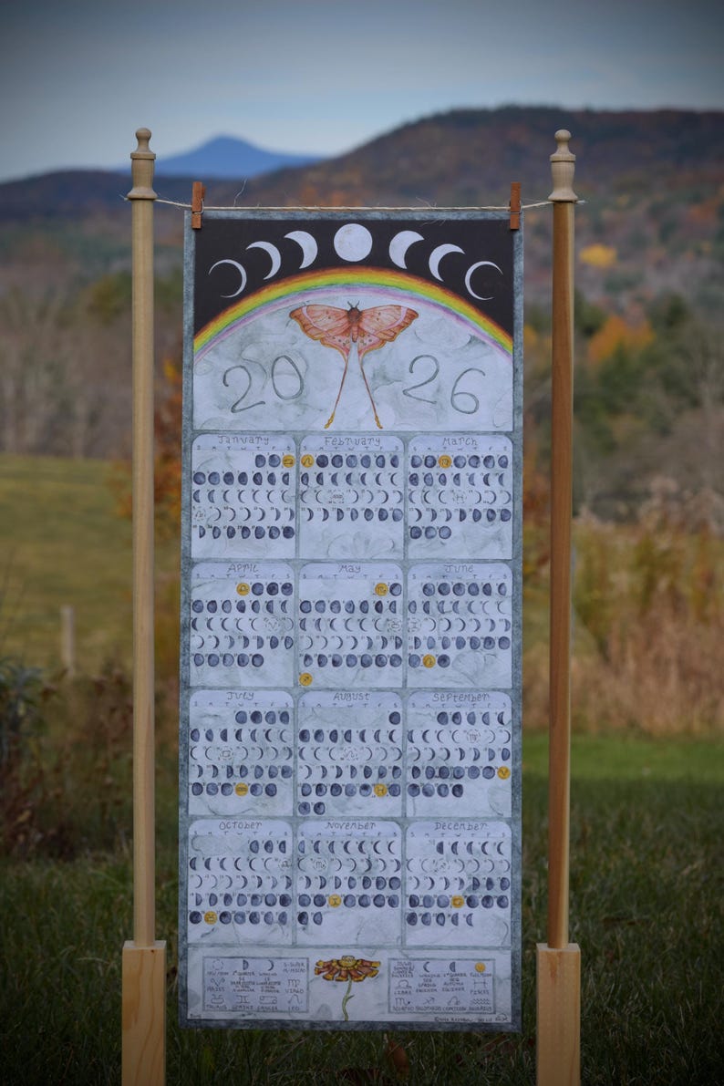 Puede incluir: Calendario de 2026 con una ilustraci&oacute;n de polilla, arco&iacute;ris y fases lunares. El calendario se muestra en un soporte de madera al aire libre, con meses y fechas dispuestas en una cuadr&iacute;cula.