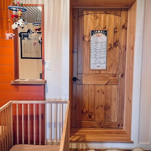 Puede incluir: Una escena de guarder&iacute;a con una cuna de madera y un colch&oacute;n marr&oacute;n, una puerta de madera con un calendario y un m&oacute;vil con figuras de animales. Las paredes est&aacute;n pintadas de naranja y blanco.