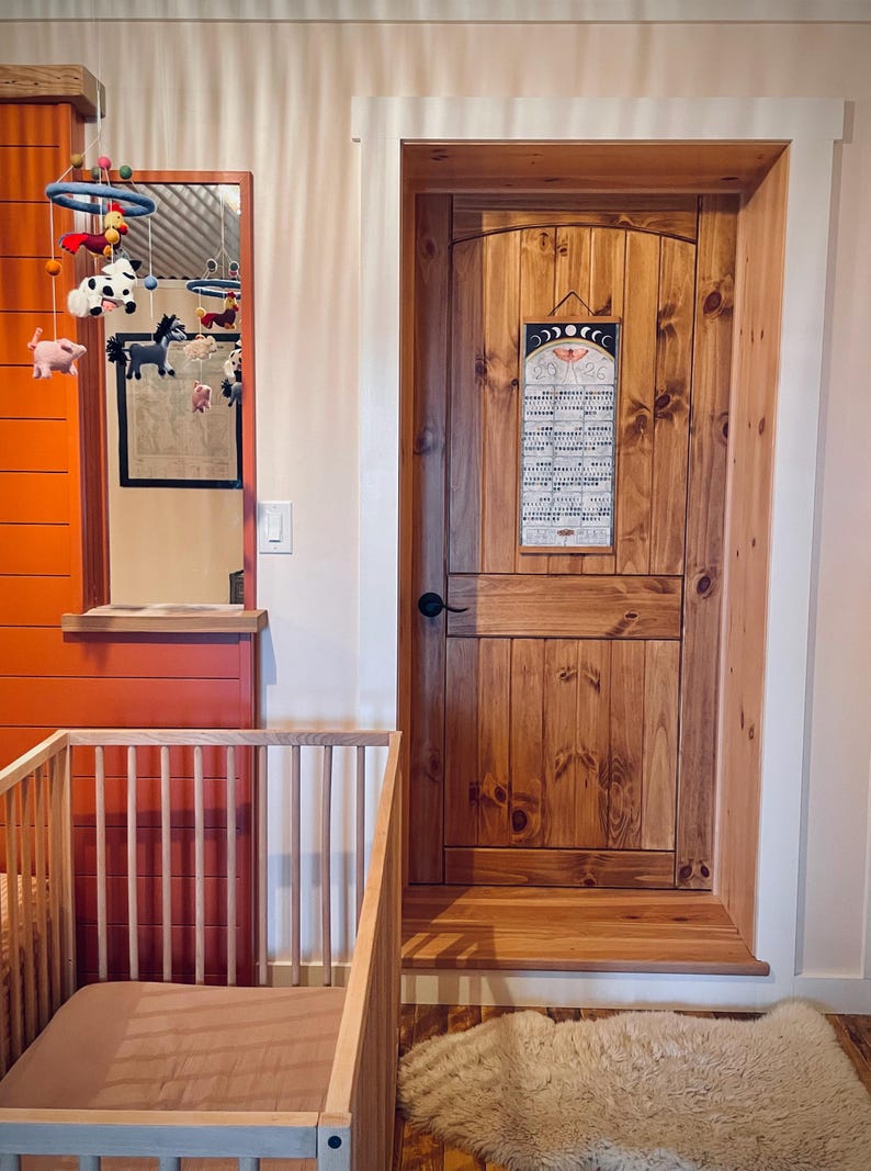 Puede incluir: Una escena de guarder&iacute;a con una cuna de madera y un colch&oacute;n marr&oacute;n, una puerta de madera con un calendario y un m&oacute;vil con figuras de animales. Las paredes est&aacute;n pintadas de naranja y blanco.