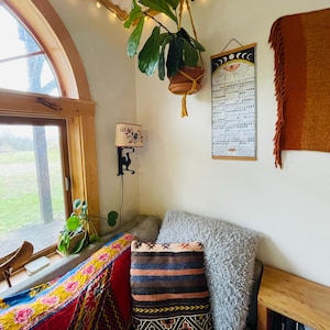 Puede incluir: Un interior acogedor con una planta colgante en una maceta marr&oacute;n, un coj&iacute;n estampado y un calendario. La habitaci&oacute;n tiene una ventana, una l&aacute;mpara peque&ntilde;a y una estanter&iacute;a de madera con libros. La iluminaci&oacute;n c&aacute;lida y un textil colorido crean un ambiente acogedor.