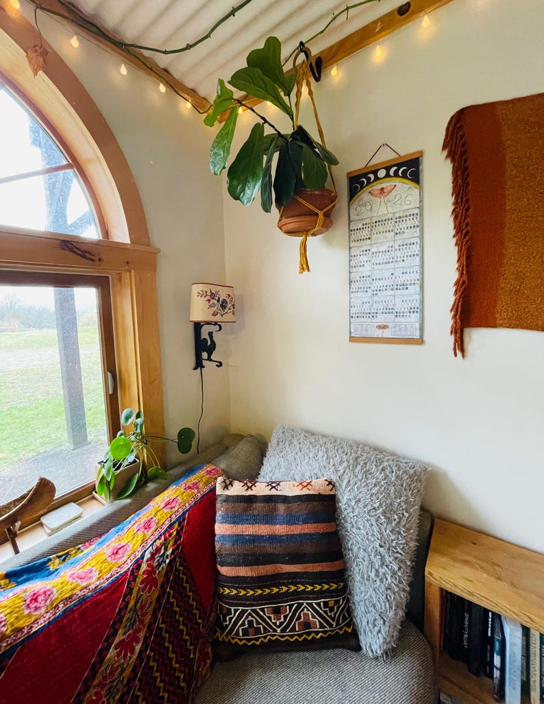 Puede incluir: Un interior acogedor con una planta colgante en una maceta marr&oacute;n, un coj&iacute;n estampado y un calendario. La habitaci&oacute;n tiene una ventana, una l&aacute;mpara peque&ntilde;a y una estanter&iacute;a de madera con libros. La iluminaci&oacute;n c&aacute;lida y un textil colorido crean un ambiente acogedor.
