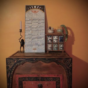 Puede incluir: Un mueble de madera con detalles pintados ornamentados, coronado por un calendario, un peque&ntilde;o cofre decorativo y un candelabro. El calendario presenta una ilustraci&oacute;n de polilla y los n&uacute;meros "20/26". El peque&ntilde;o cofre tiene seis baldosas decorativas.