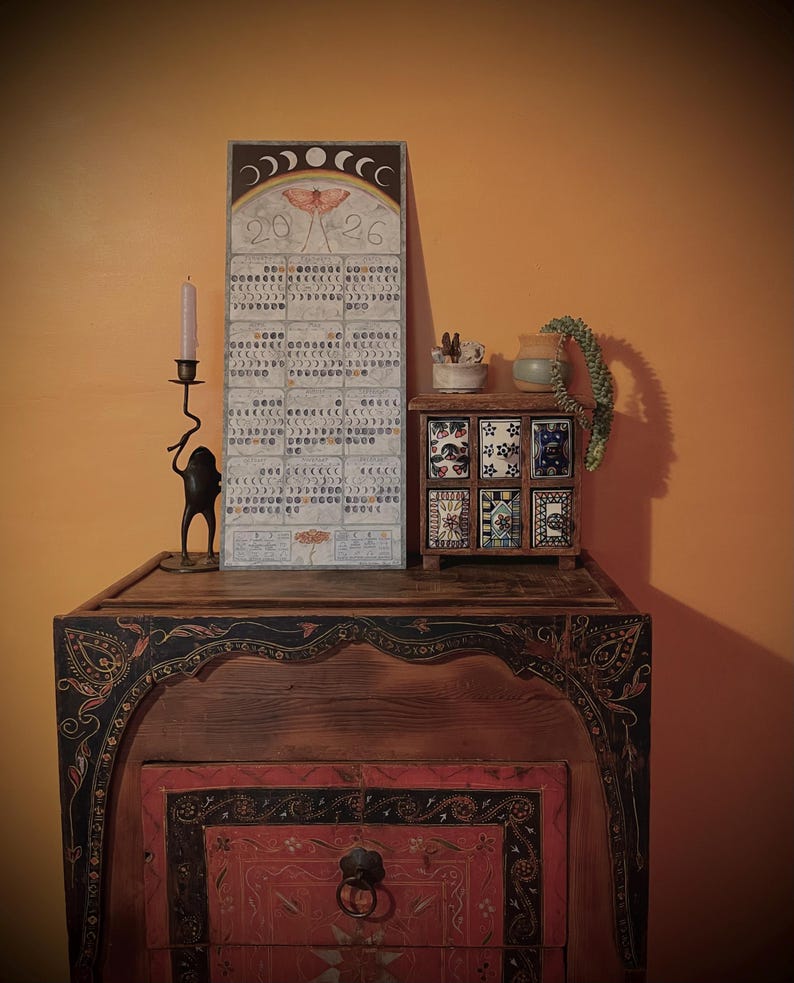 Puede incluir: Un mueble de madera con detalles pintados ornamentados, coronado por un calendario, un peque&ntilde;o cofre decorativo y un candelabro. El calendario presenta una ilustraci&oacute;n de polilla y los n&uacute;meros "20/26". El peque&ntilde;o cofre tiene seis baldosas decorativas.