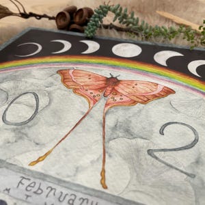 Puede incluir: Ilustraci&oacute;n en acuarela de una p&aacute;gina de calendario con una polilla de alas largas y fluidas, un arco&iacute;ris y fases lunares. El a&ntilde;o "2020" es visible, junto con el mes de febrero. La paleta de colores incluye tonos de naranja, rosa y gris.