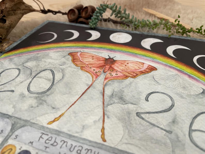 Puede incluir: Ilustraci&oacute;n en acuarela de una p&aacute;gina de calendario con una polilla de alas largas y fluidas, un arco&iacute;ris y fases lunares. El a&ntilde;o "2020" es visible, junto con el mes de febrero. La paleta de colores incluye tonos de naranja, rosa y gris.